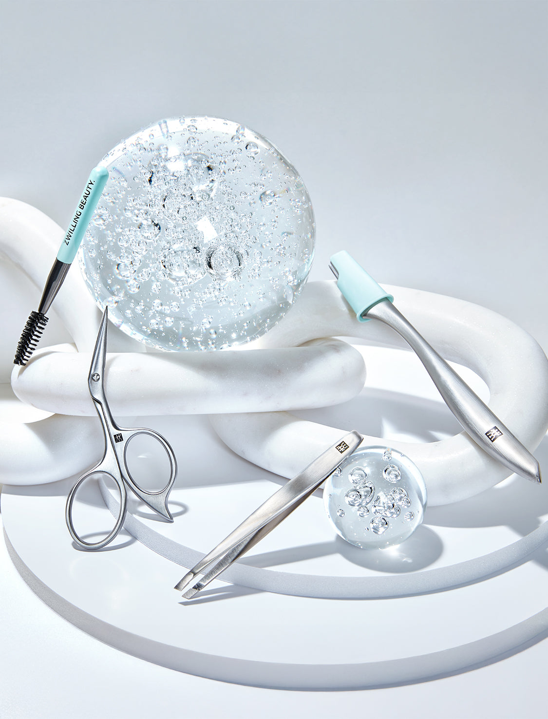 Zwilling-Schönheitswerkzeuge arrangiert auf weißen Marmobjekten mit Glaskugeln vor minimalistischem, weißem Hintergrund.