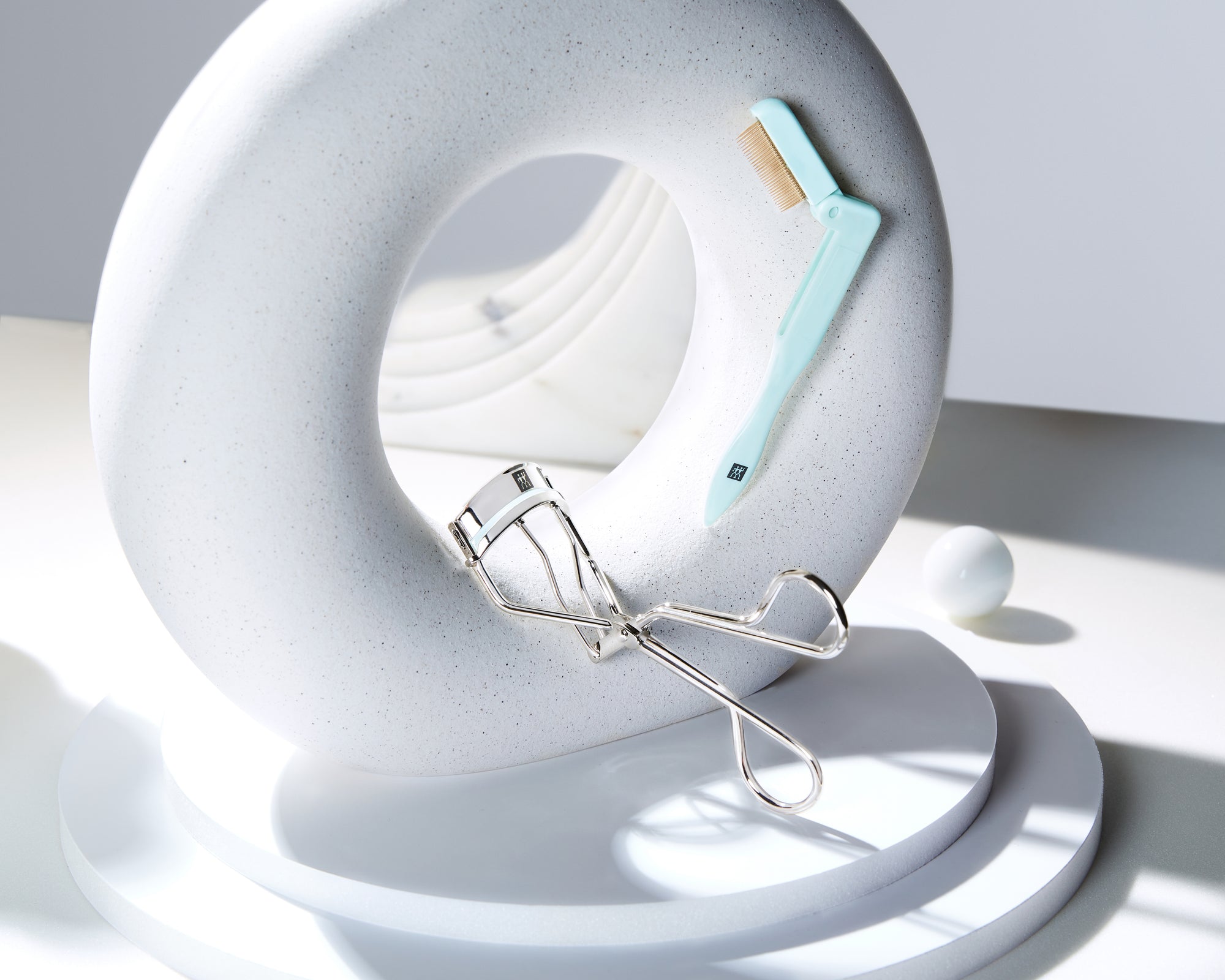 Darstellung der ZWILLING Premium Wimpernzange und dem Premium Wimpernkamm umgeben von artifiziellen Steinobjekten.