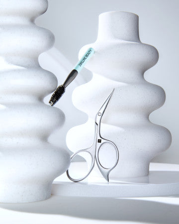 Darstellung einer ZWILLING Premium Augenbrauenschere aus rostfreiem Edelstahl und Augenbrauenbürste inmitten von kunstvoll geformten Keramikobjekten.