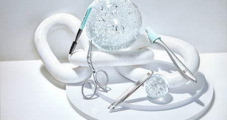 ZWILLING Augenbrauen Werkzeuge in Silber auf Glaskugeln und Marmorobjekten drapiert.